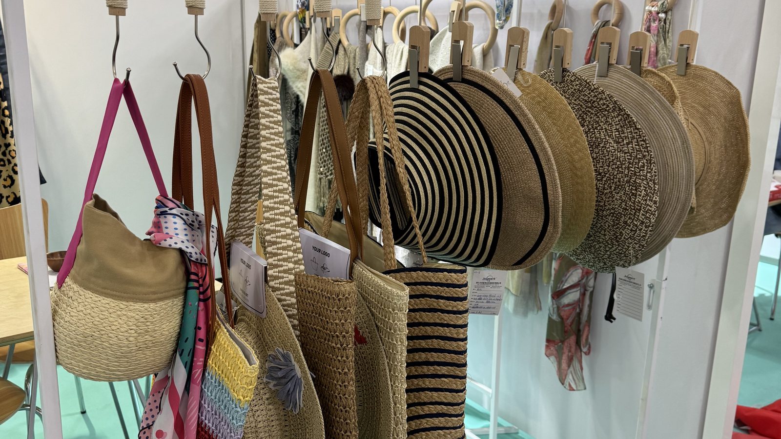 A row of straw hats and woven bags on a white display rack over mint-green carpet at our Texworld NYC 2026 booth.