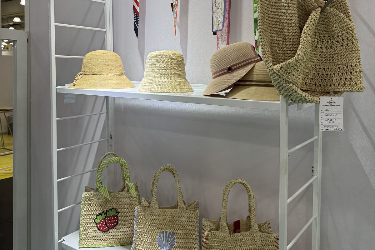 Shelf display of hats, mini woven totes with embroidered fruit and shell motifs, and ribbon trims.